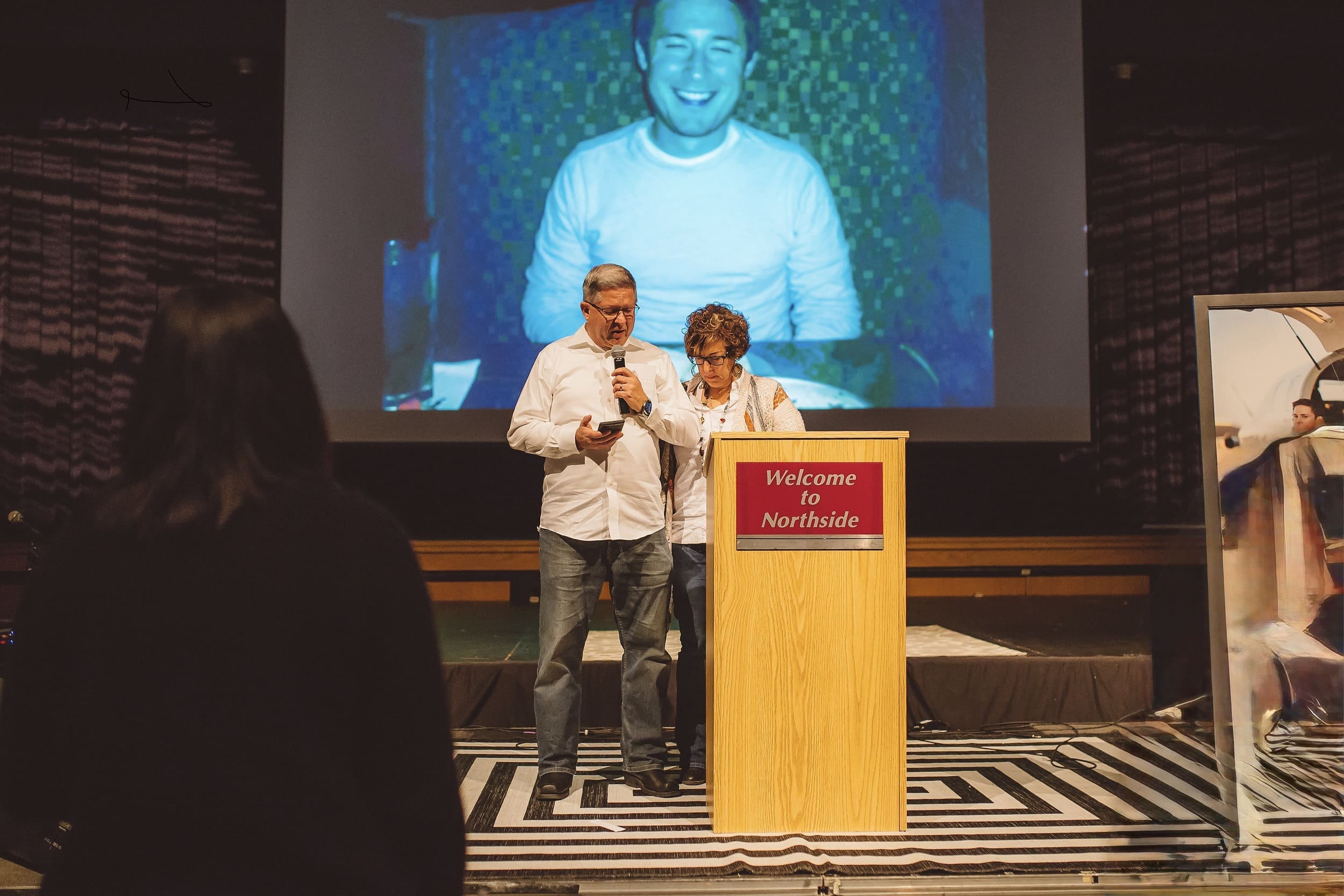 Grieving parents praying and standing in front of a projected photo of the son they lost